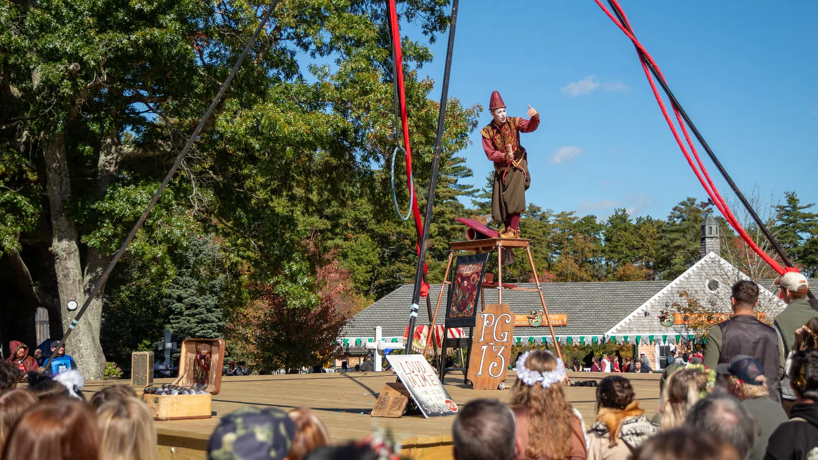 Street performer in medieval costume entertaining a crowd on an outdoor stage under blue sky.