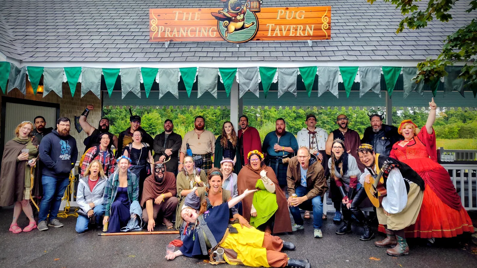 Group of people in colorful medieval costumes posing outside The Prancing Pug Tavern with green bunting overhead.
