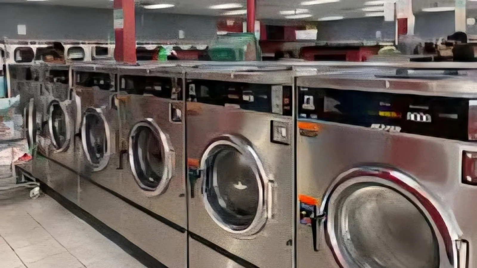 Row of industrial stainless steel washing machines in a brightly lit laundromat interior with tiled floor.