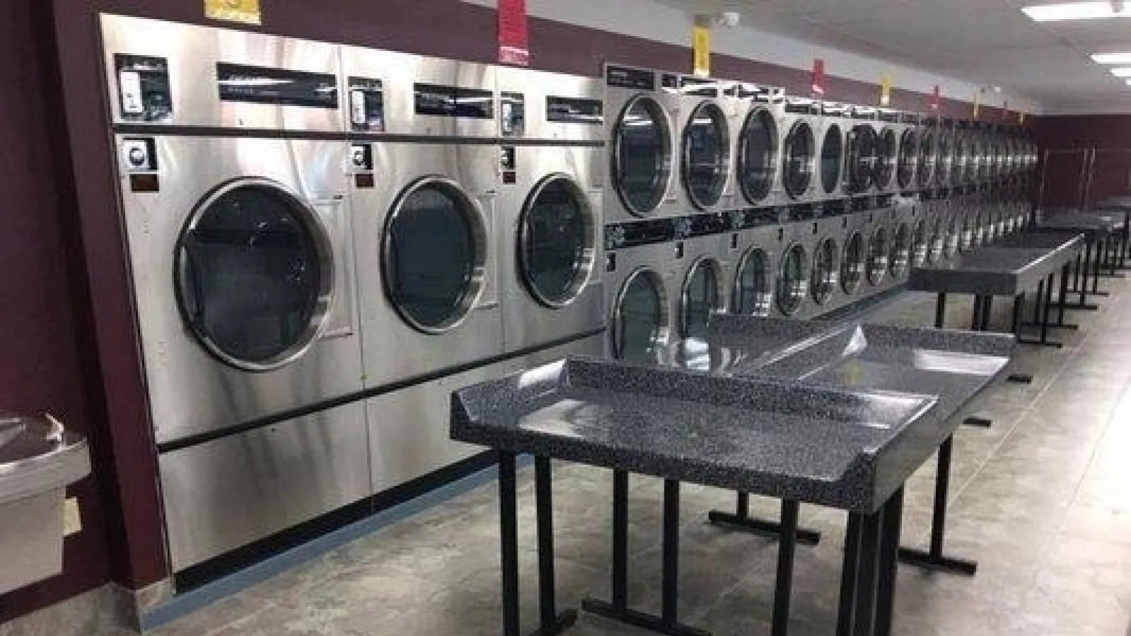 Modern laundromat with rows of stainless steel washers and dryers and folding tables on tiled floor