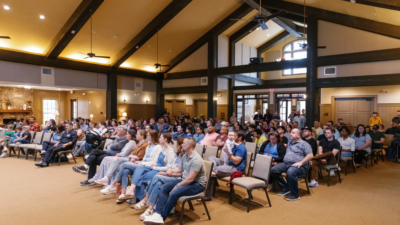 Large audience seated looking towards the front in a spacious, modern conference room with high ceilings and exposed beams.