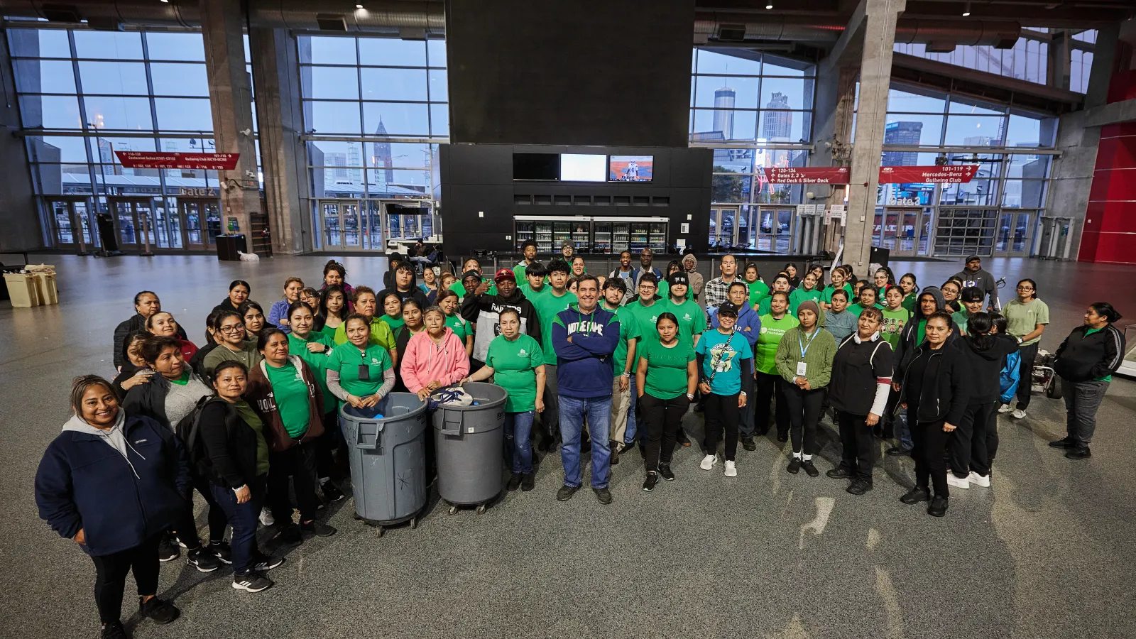 Large diverse group of people posing indoors with recycling bins in a spacious hall with large windows.
