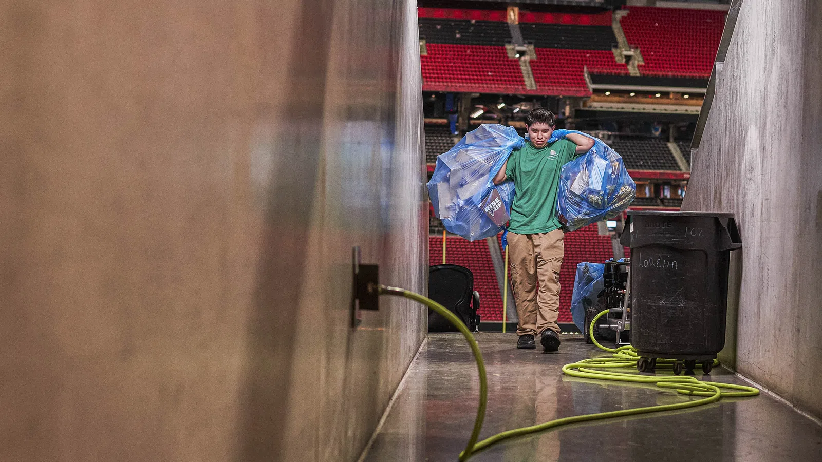 Person carrying large bags of recyclable plastic bottles through a stadium corridor with equipment and hoses present.