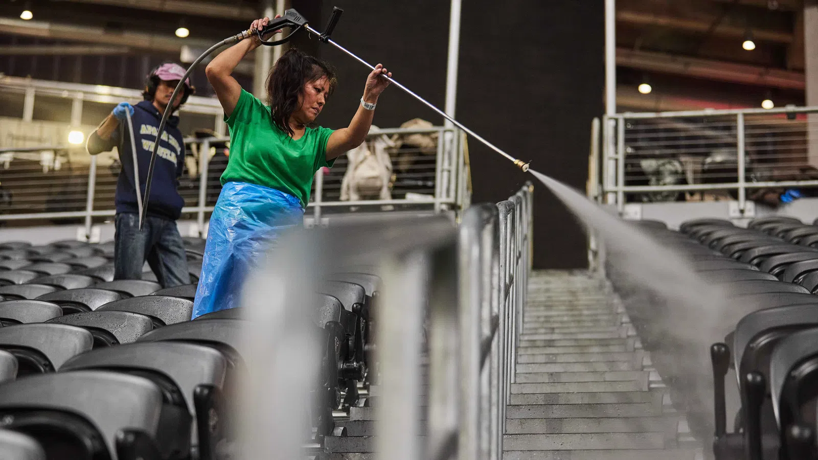 Woman power washing stadium seats and stairs while another person cleans seats in the background.