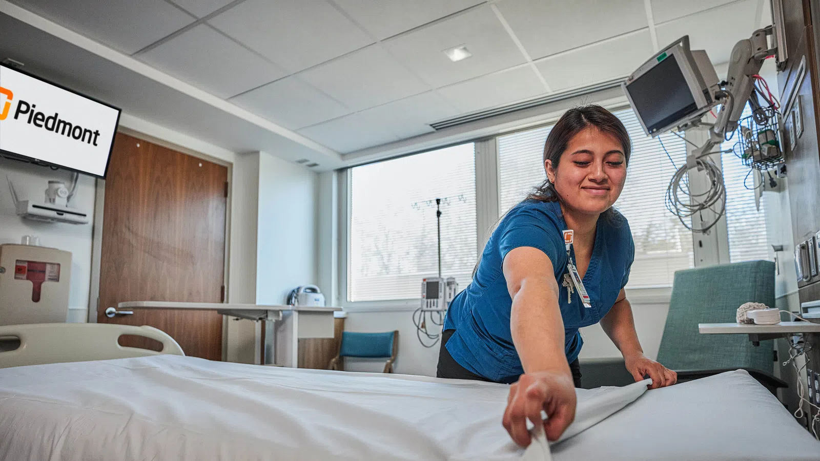 Healthcare worker in blue scrubs making a hospital bed in a bright patient room with Piedmont logo on screen.