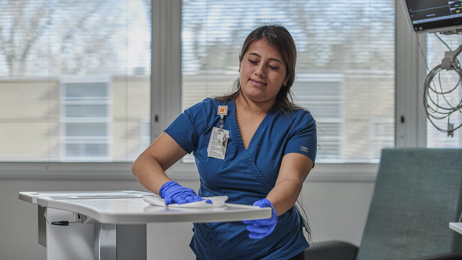 Healthcare worker in blue scrubs and gloves cleaning hospital room surface near medical equipment.