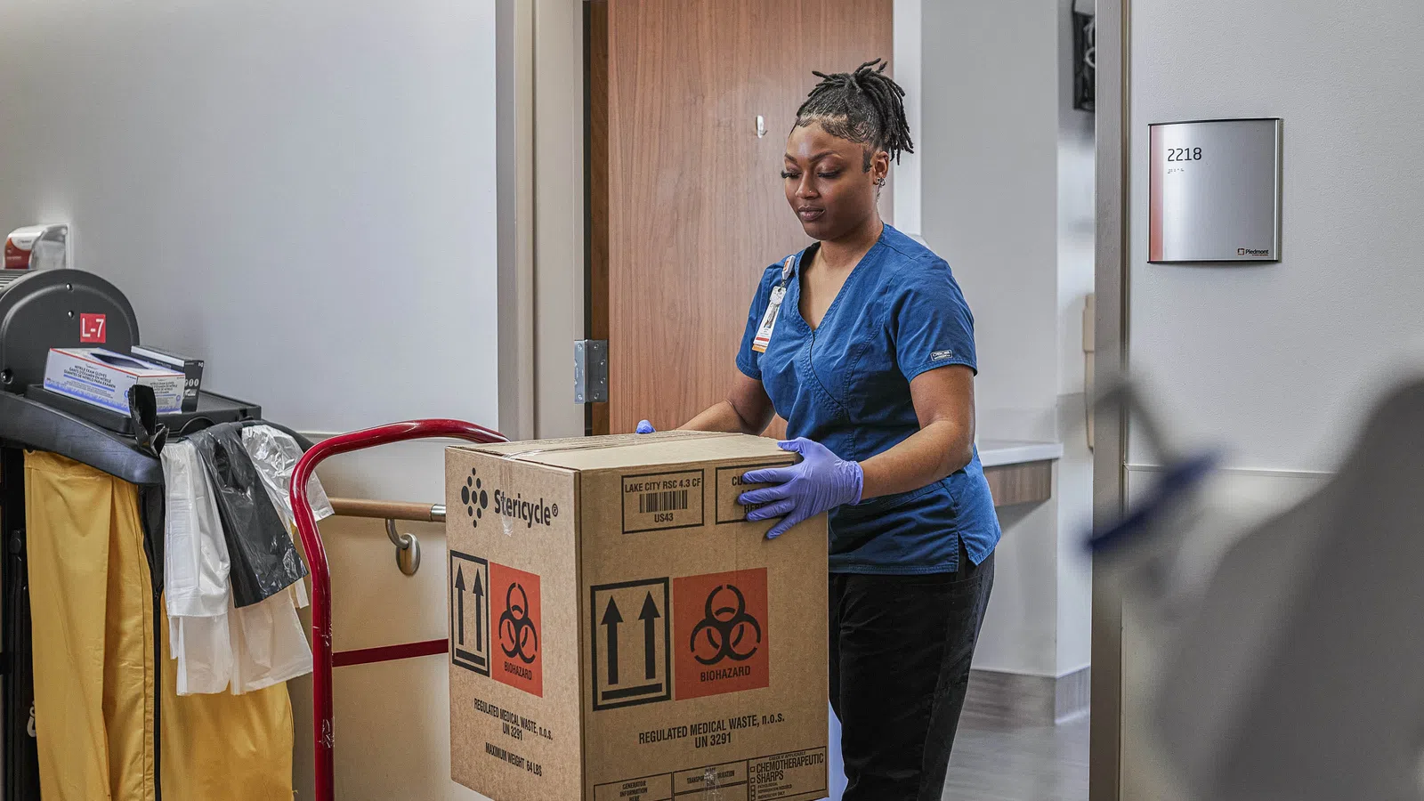 Healthcare worker in scrubs handling a biohazard waste box inside a medical facility hallway.