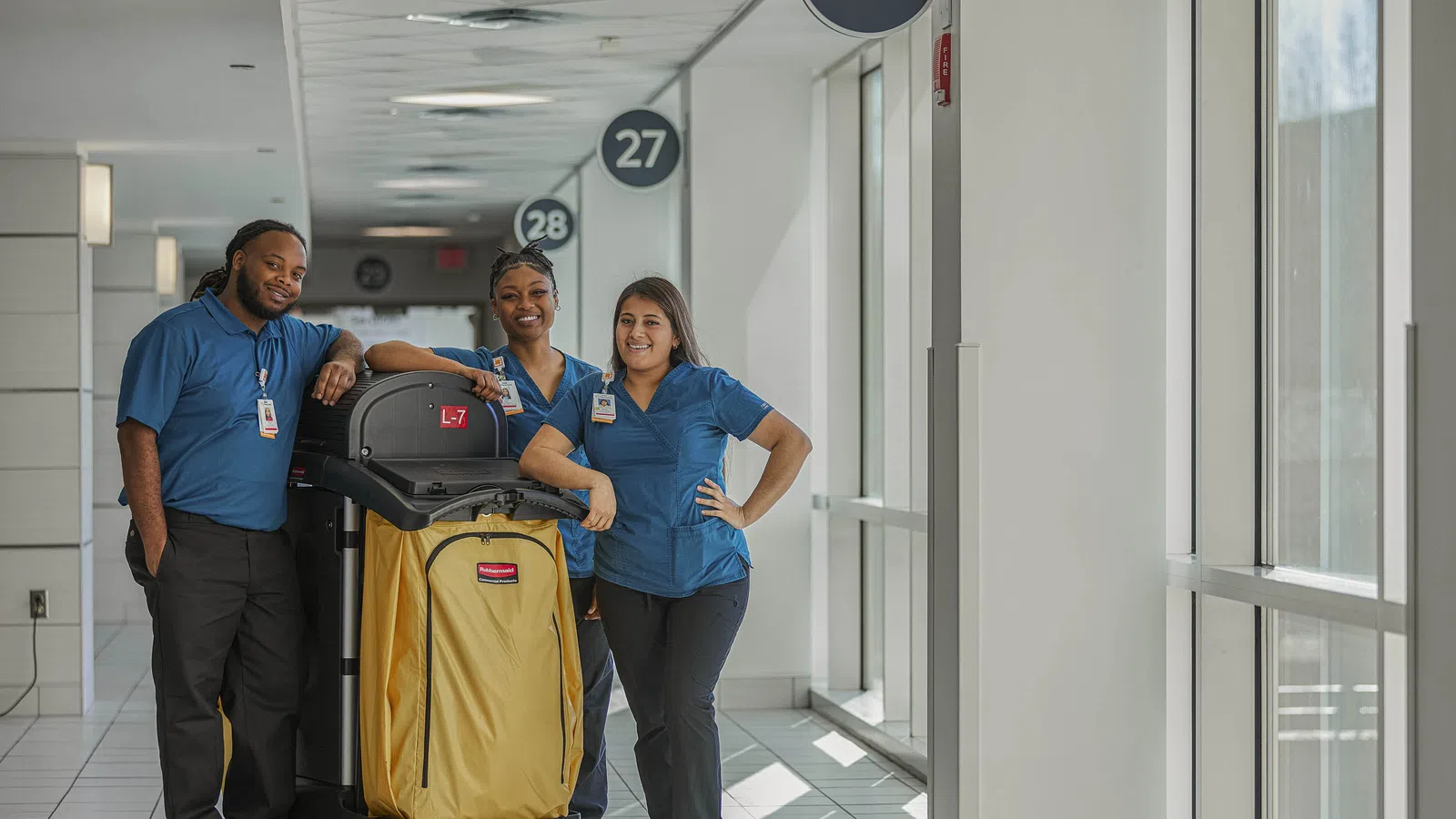 Three cleaning staff in blue uniforms standing with a yellow cart in a bright hallway near numbered doors.