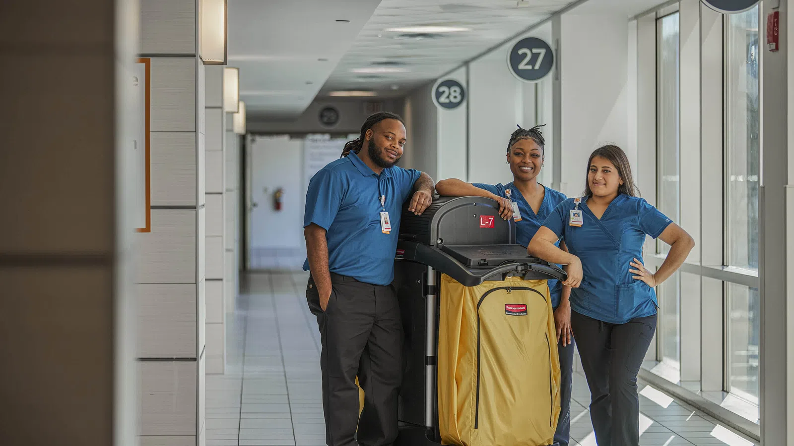 Three janitors in blue uniforms standing with a yellow cleaning cart in a bright hallway with numbered signs.