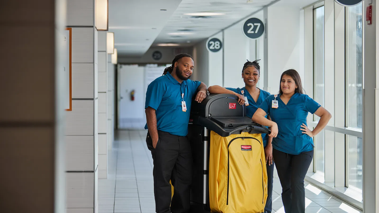 Three cleaning staff in blue uniforms posing with a large yellow housekeeping cart in a bright hallway.