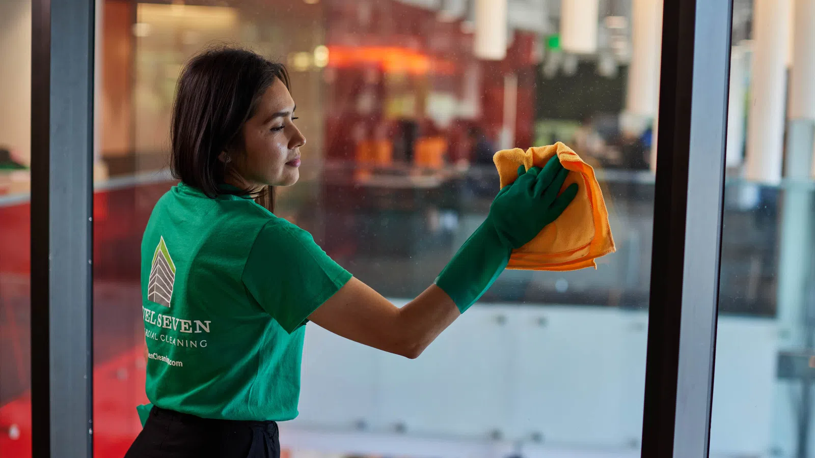 Professional cleaner in green gloves wiping a glass window with an orange cloth in an office building.