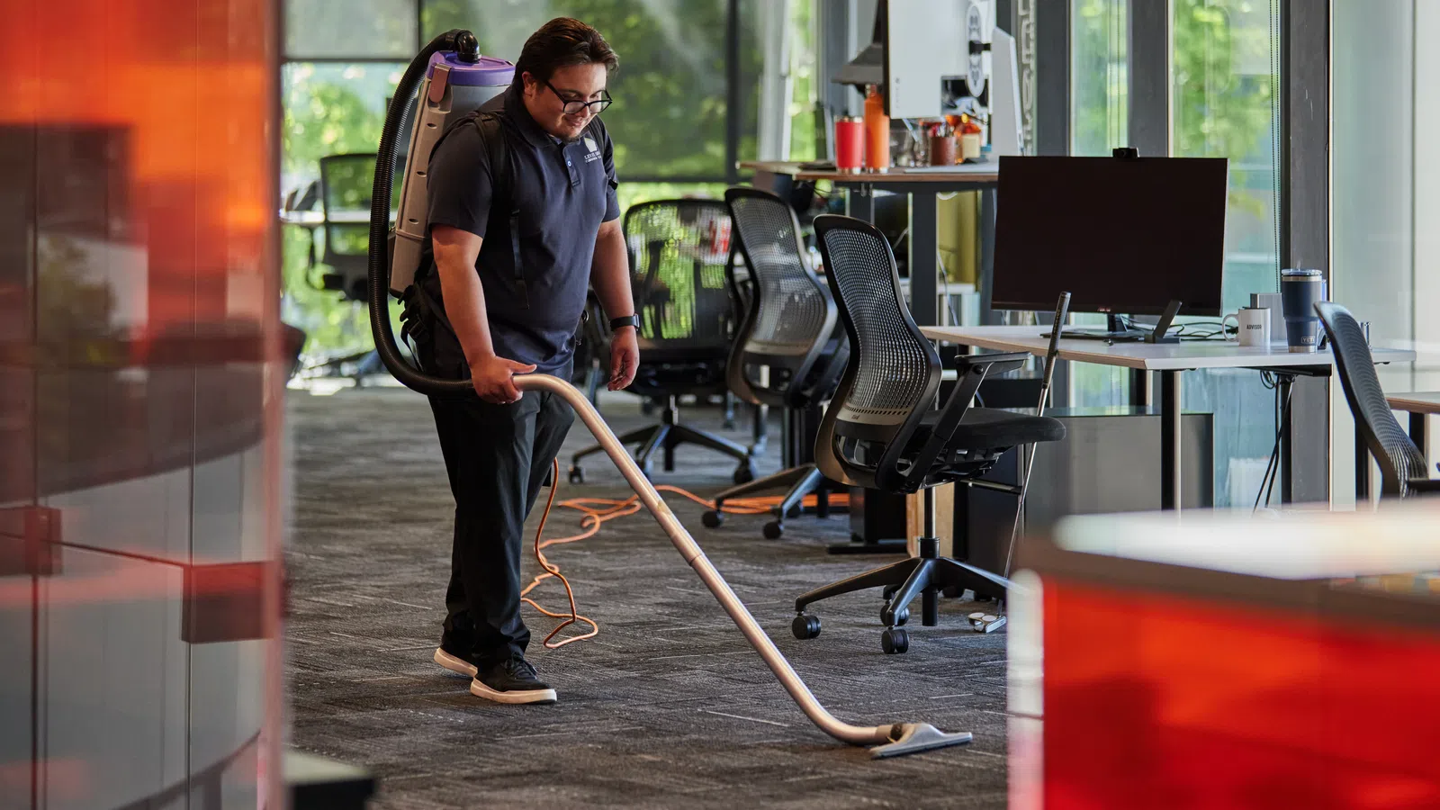 Janitor vacuuming carpet in a modern office with glass walls and ergonomic chairs during daytime.