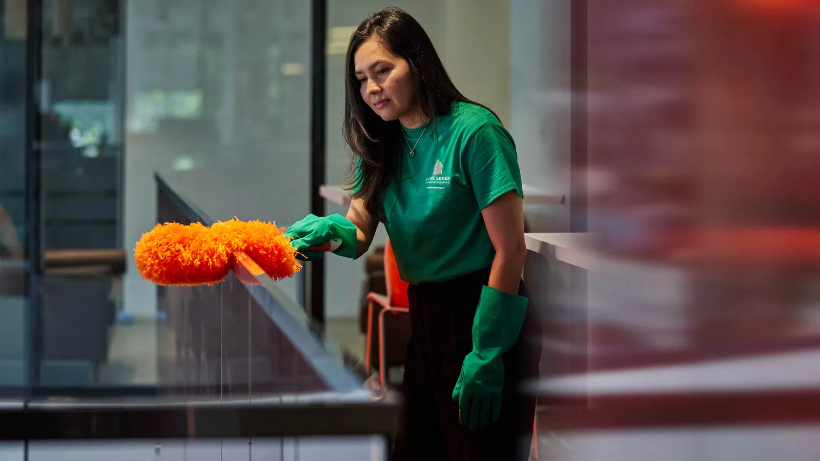 Woman wearing green gloves dusting a glass railing with an orange duster in a modern indoor space.