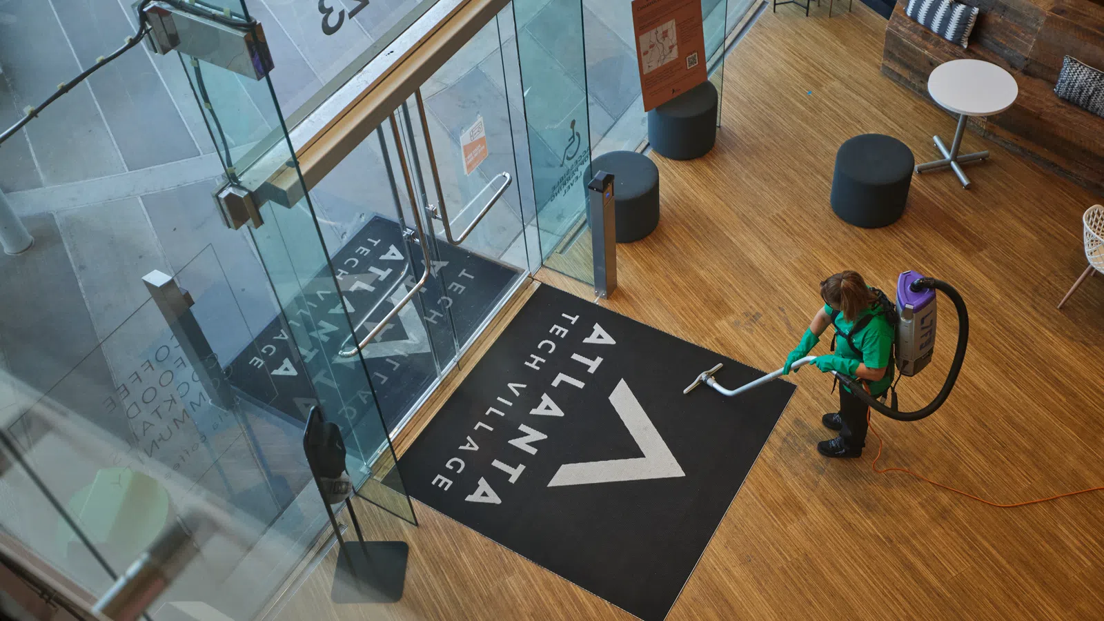 Person vacuuming wooden floor near glass entrance with Atlanta Tech Village logo mat inside building lobby
