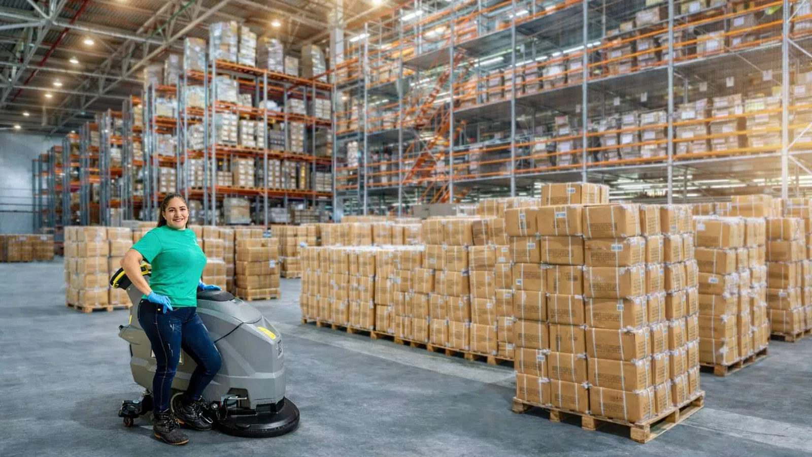 Woman operating a Kärcher Professional floor cleaning machine in a large industrial warehouse.