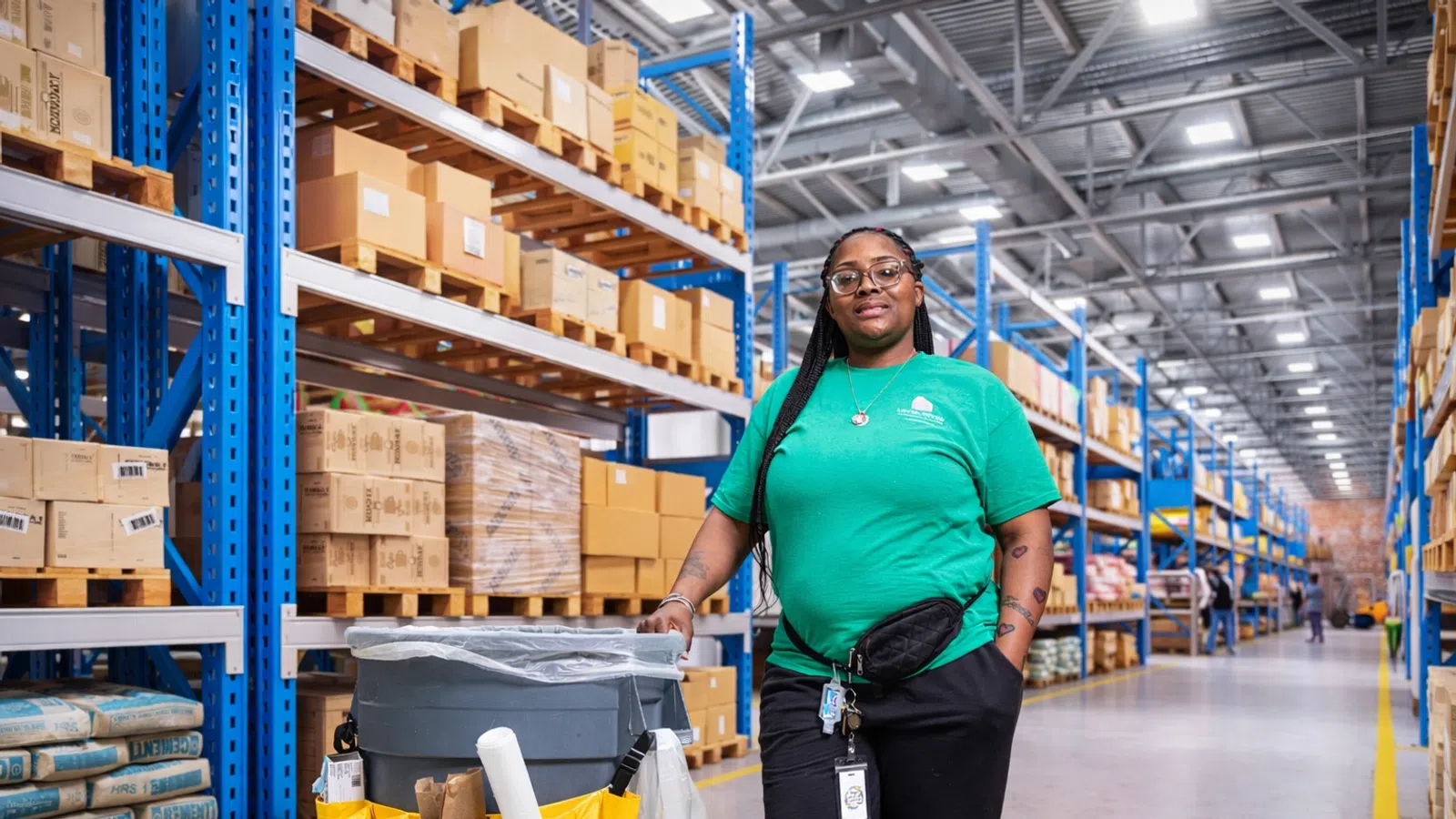 Warehouse worker in green shirt standing next to cleaning cart in aisle with shelves of boxes and supplies.