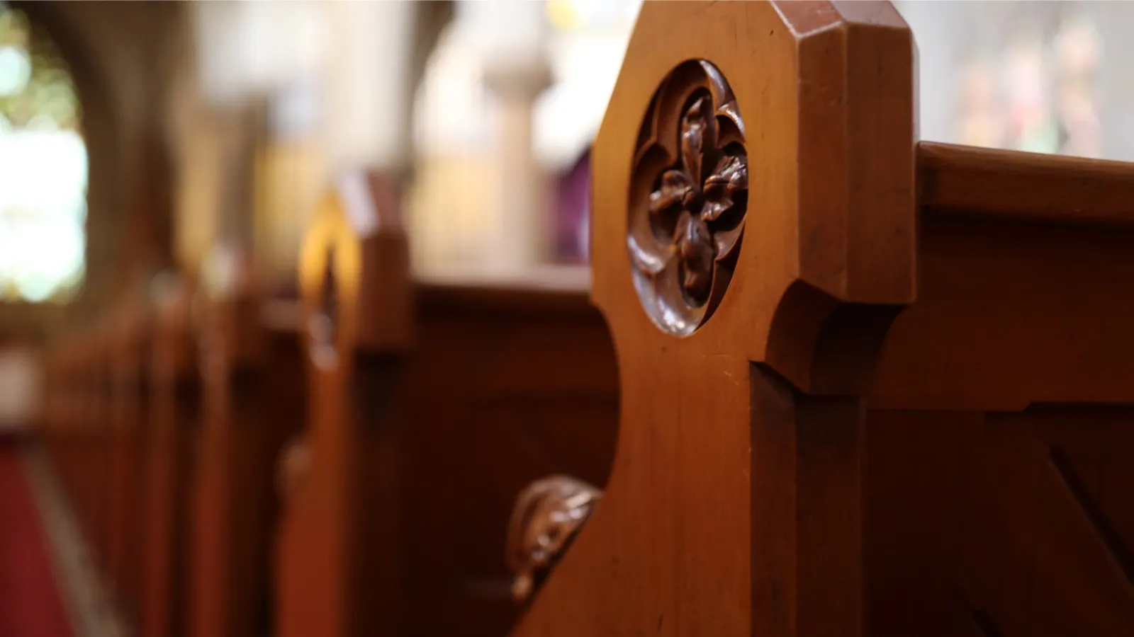 Close-up of carved wooden pews inside a church with stained glass windows in soft background light