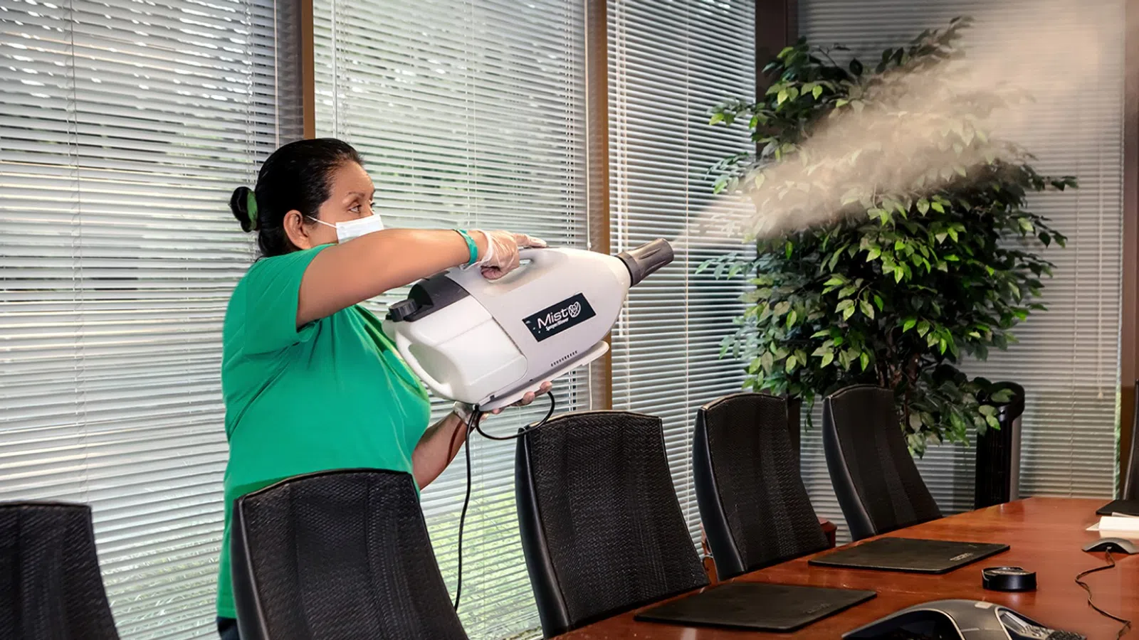 Person wearing mask spraying disinfectant fog in a modern office conference room with glass windows and chairs.