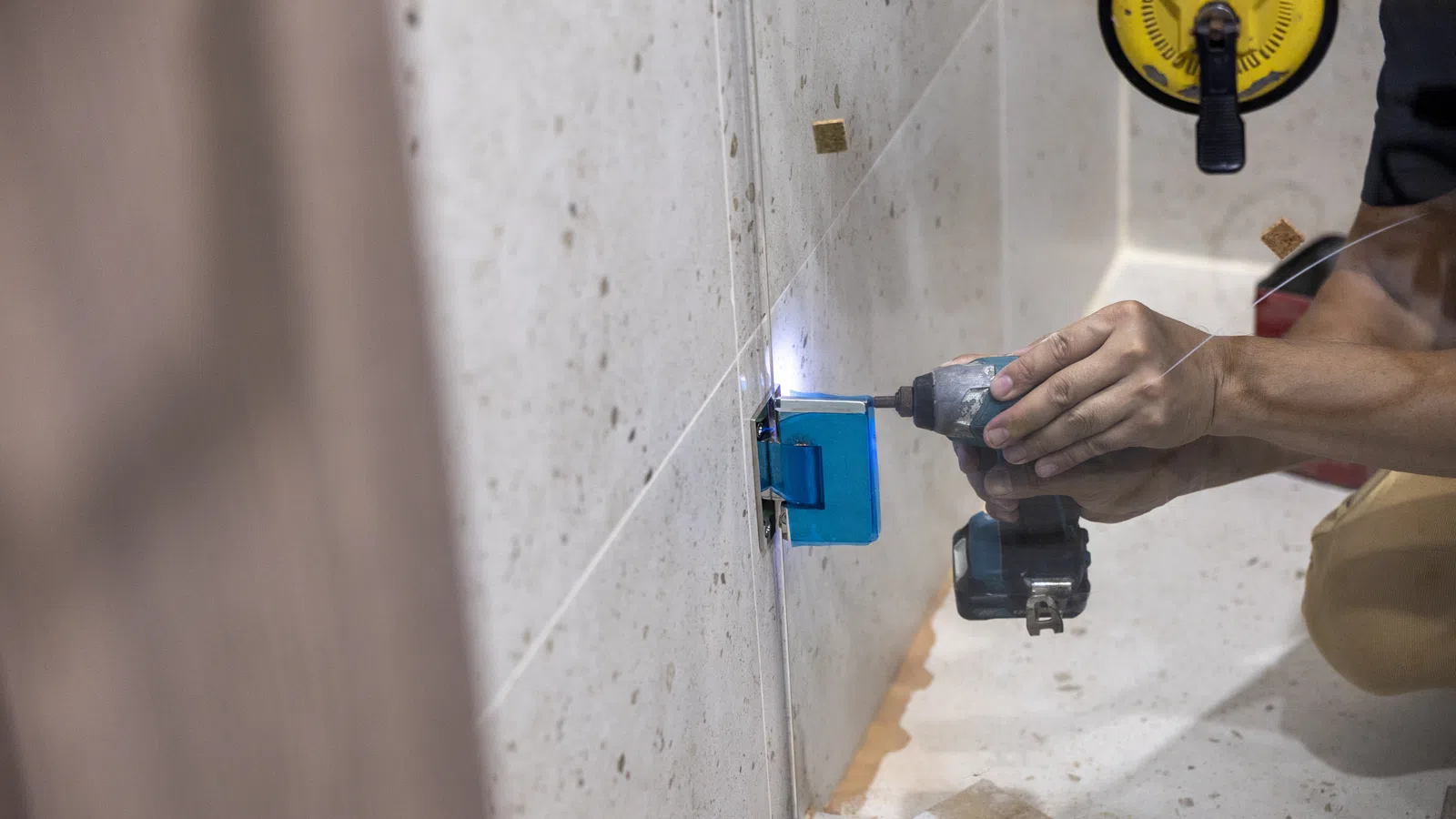 Person using a cordless drill to install a glass shower door in a bathroom under construction.