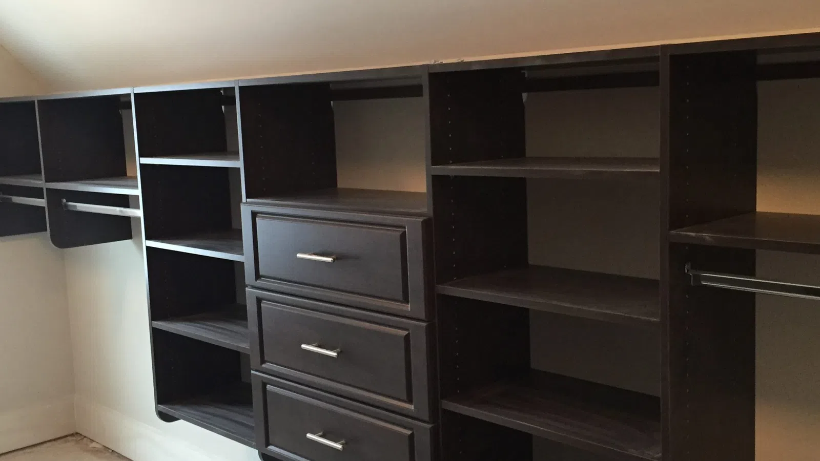 Custom dark wood closet organizer with shelves, hanging rods, and three drawers in a walk-in closet.