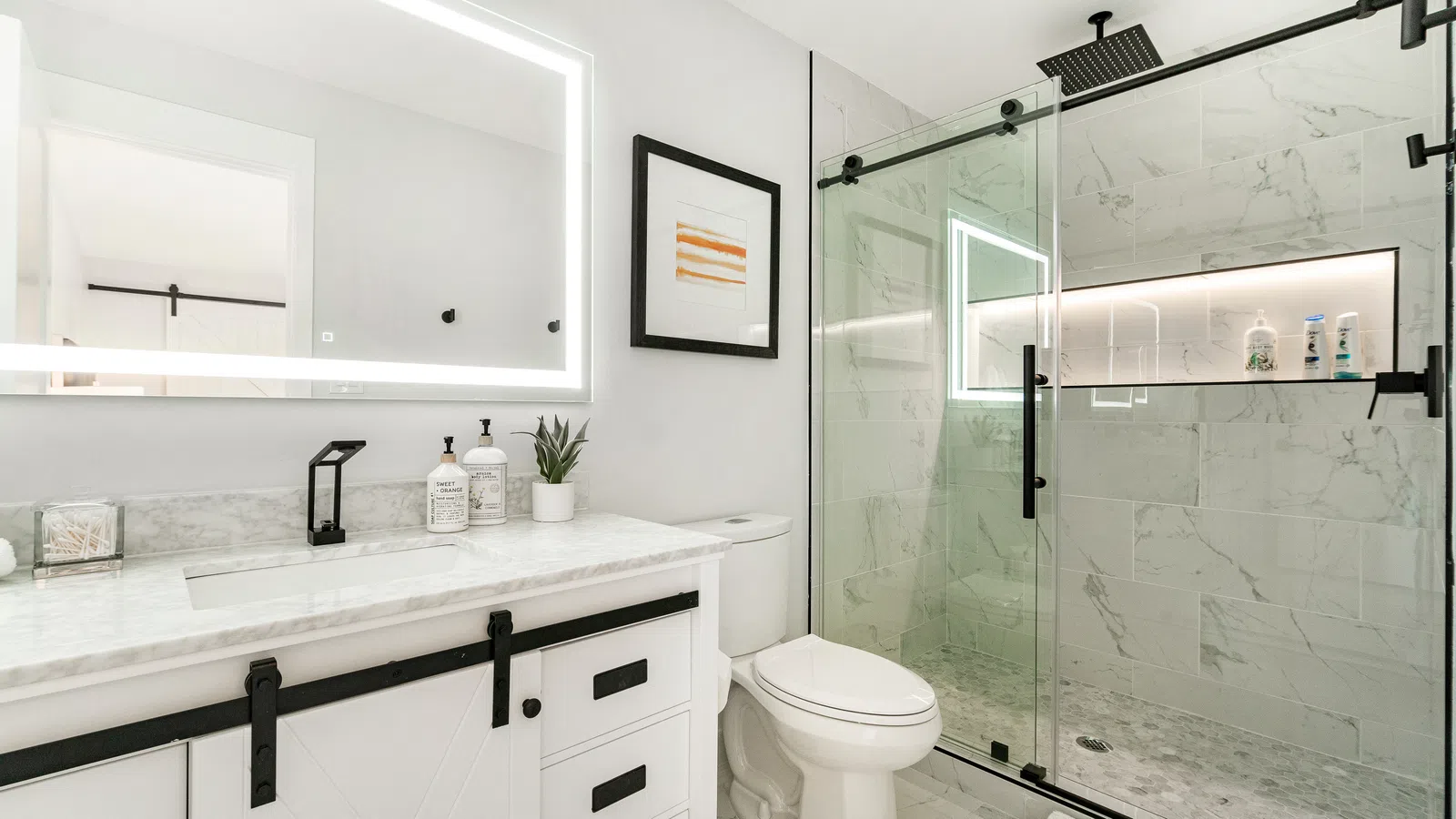 Modern white bathroom with illuminated mirror, marble countertop, black fixtures, and glass shower enclosure