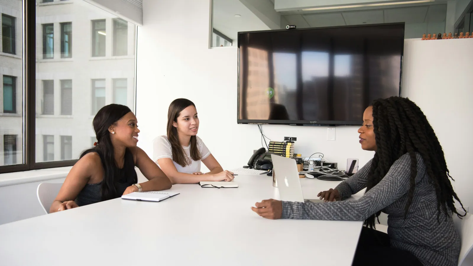 Three women engaged in a meeting around a white conference table with laptop and notebooks in a modern office.
