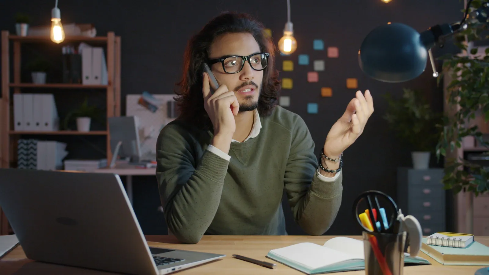 Young man with glasses talking on phone at desk with laptop and notebook in cozy workspace