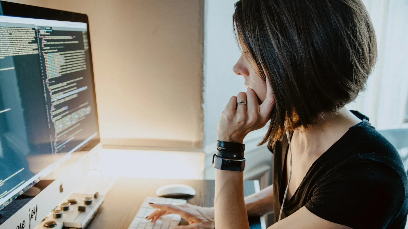 Woman focused on coding on a large desktop monitor at a wooden desk in a bright room