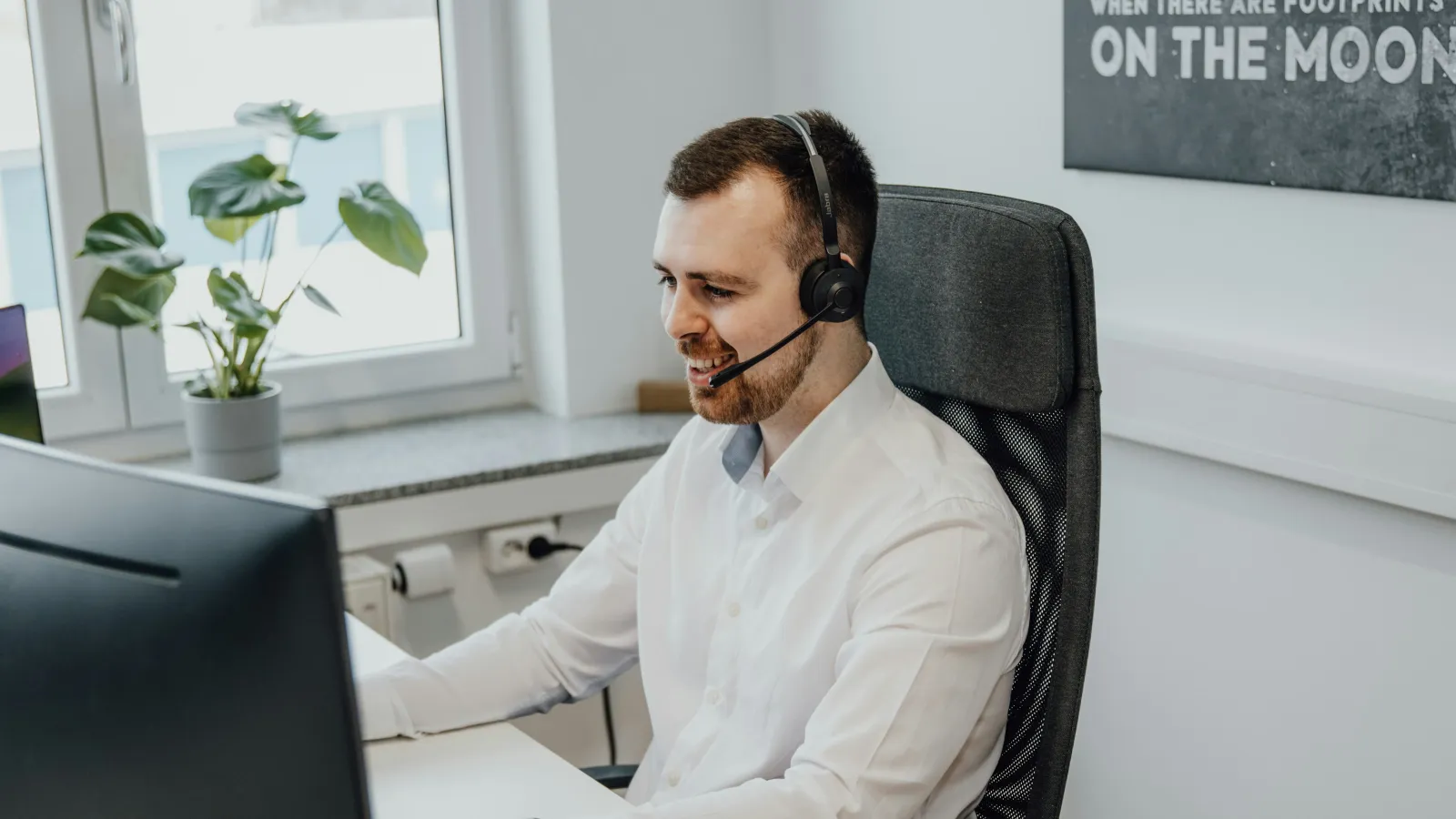 Smiling man wearing headset working on computer in modern office with plant and motivational poster