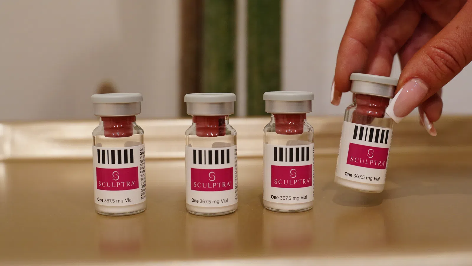 Four Sculptra vials with red caps, one held by a hand with manicured nails, placed on a reflective surface.