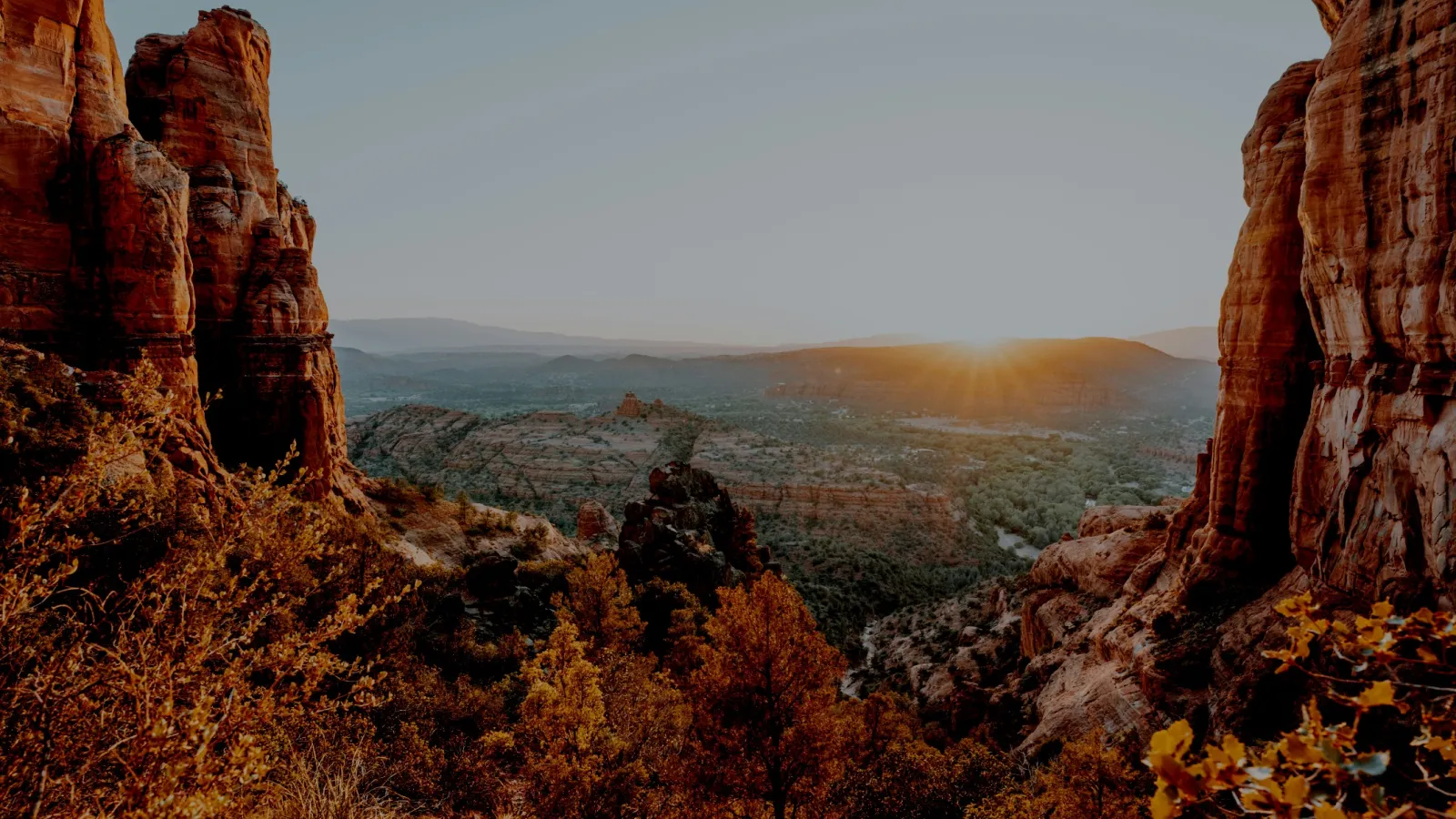 Sunrise over rocky red cliffs and autumn trees in a vast desert landscape with distant mountains.