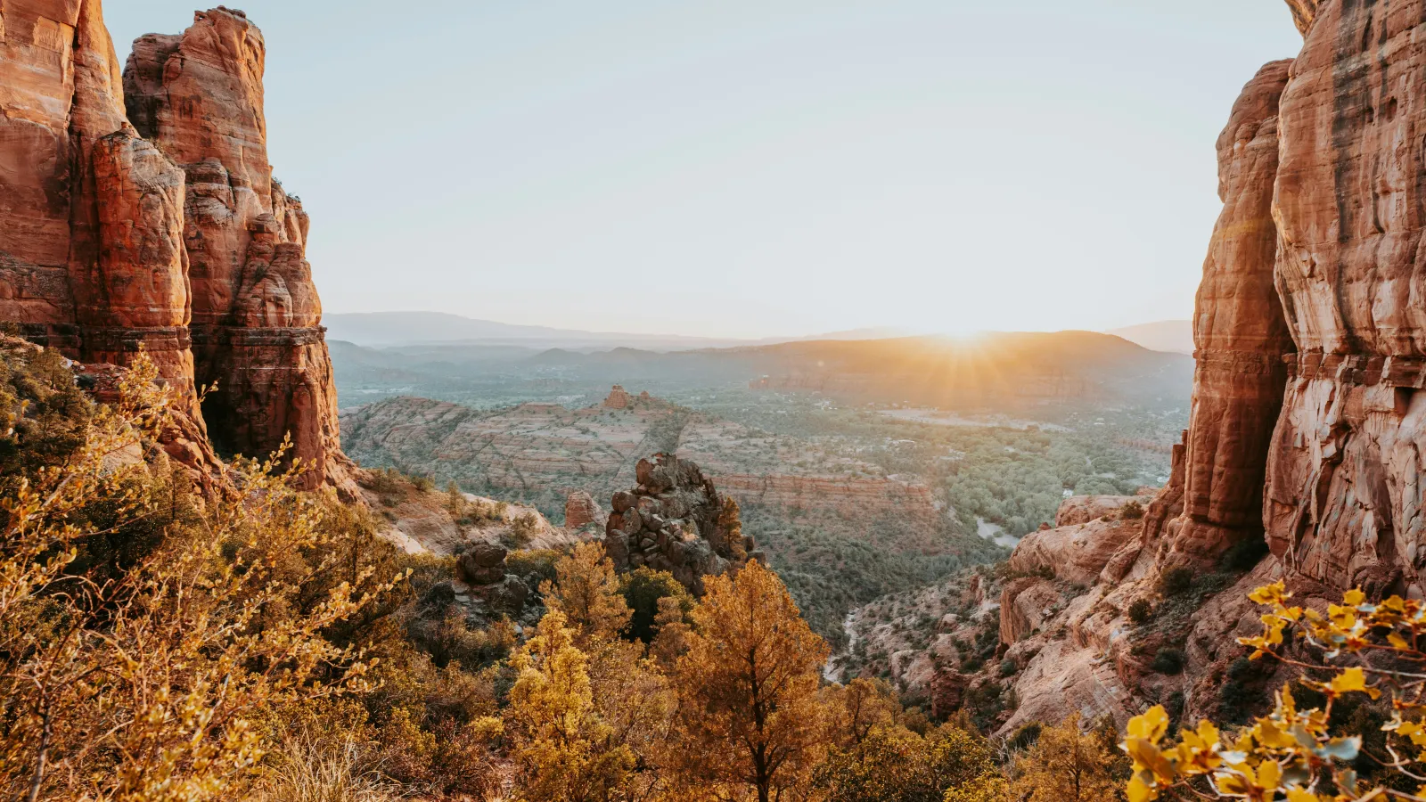 Sunrise over rocky red cliffs and autumn trees in a vast desert landscape with distant mountains.