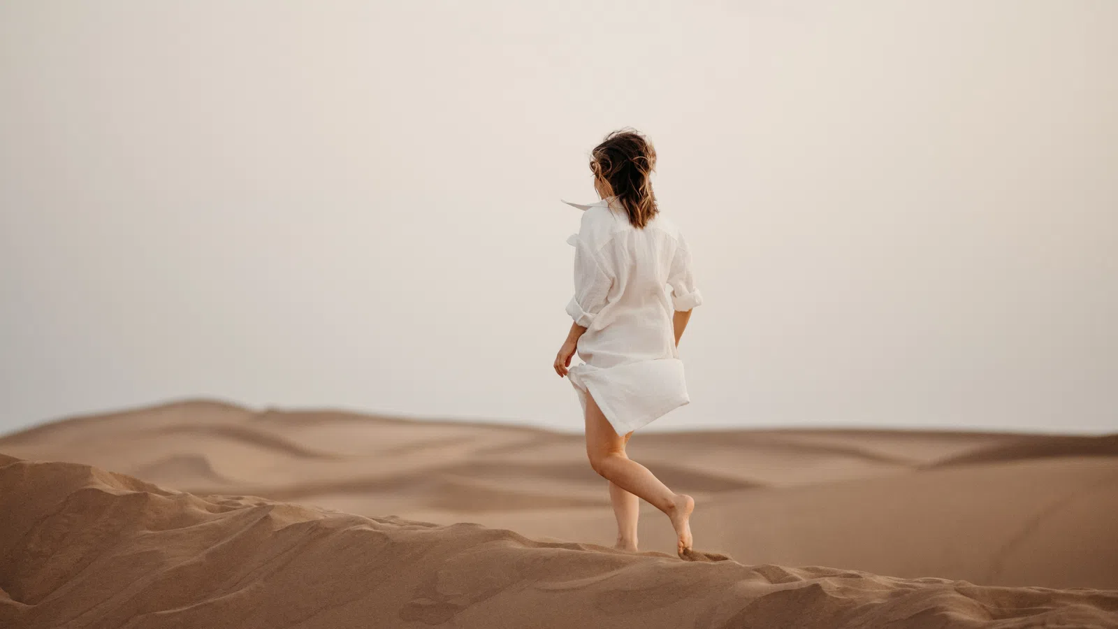 Woman in white shirt walking barefoot across sandy desert dunes at sunset with soft lighting.
