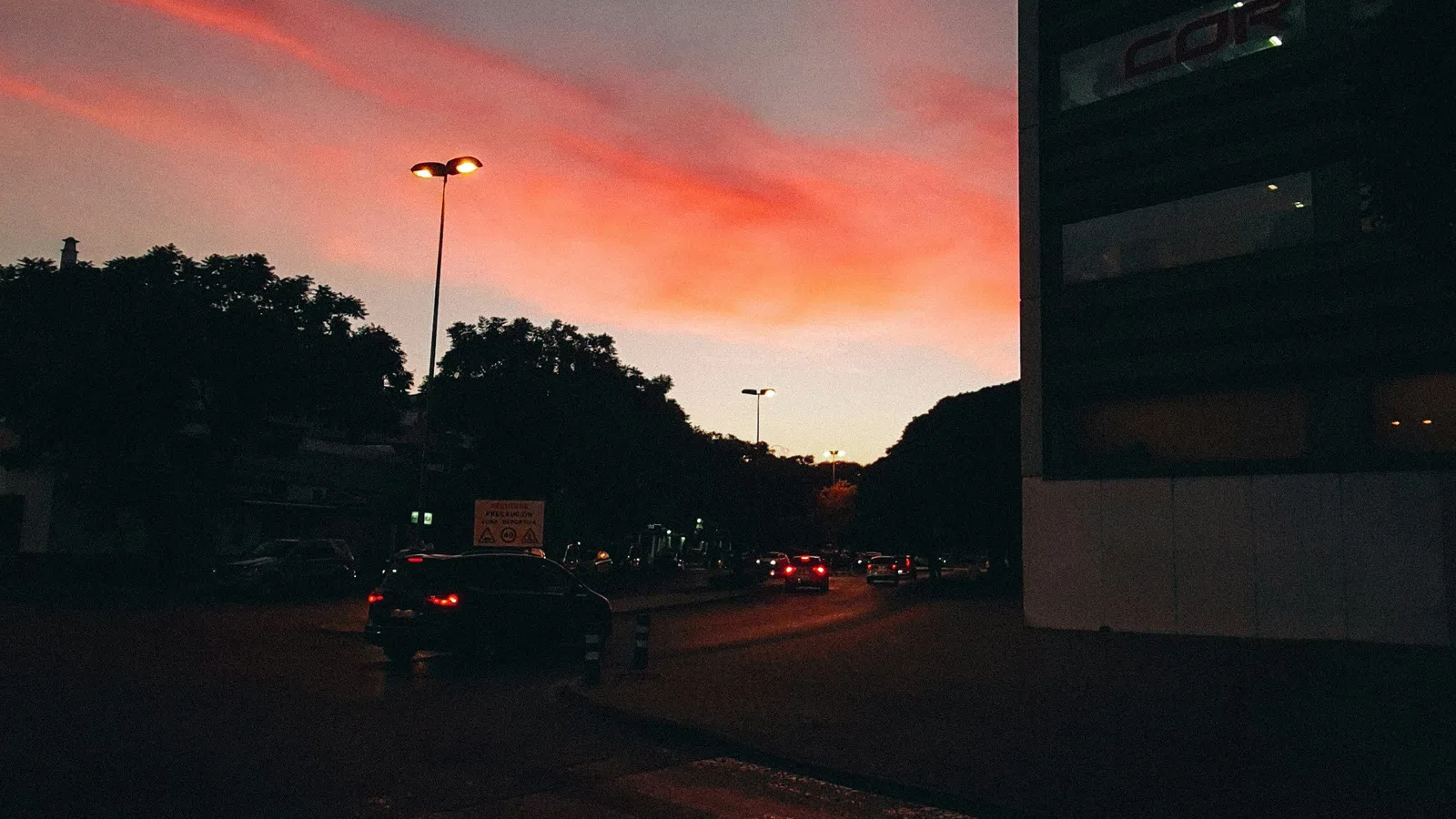 Urban street scene at dusk with dark silhouettes, cars, streetlights, and orange-pink clouds in the sky.