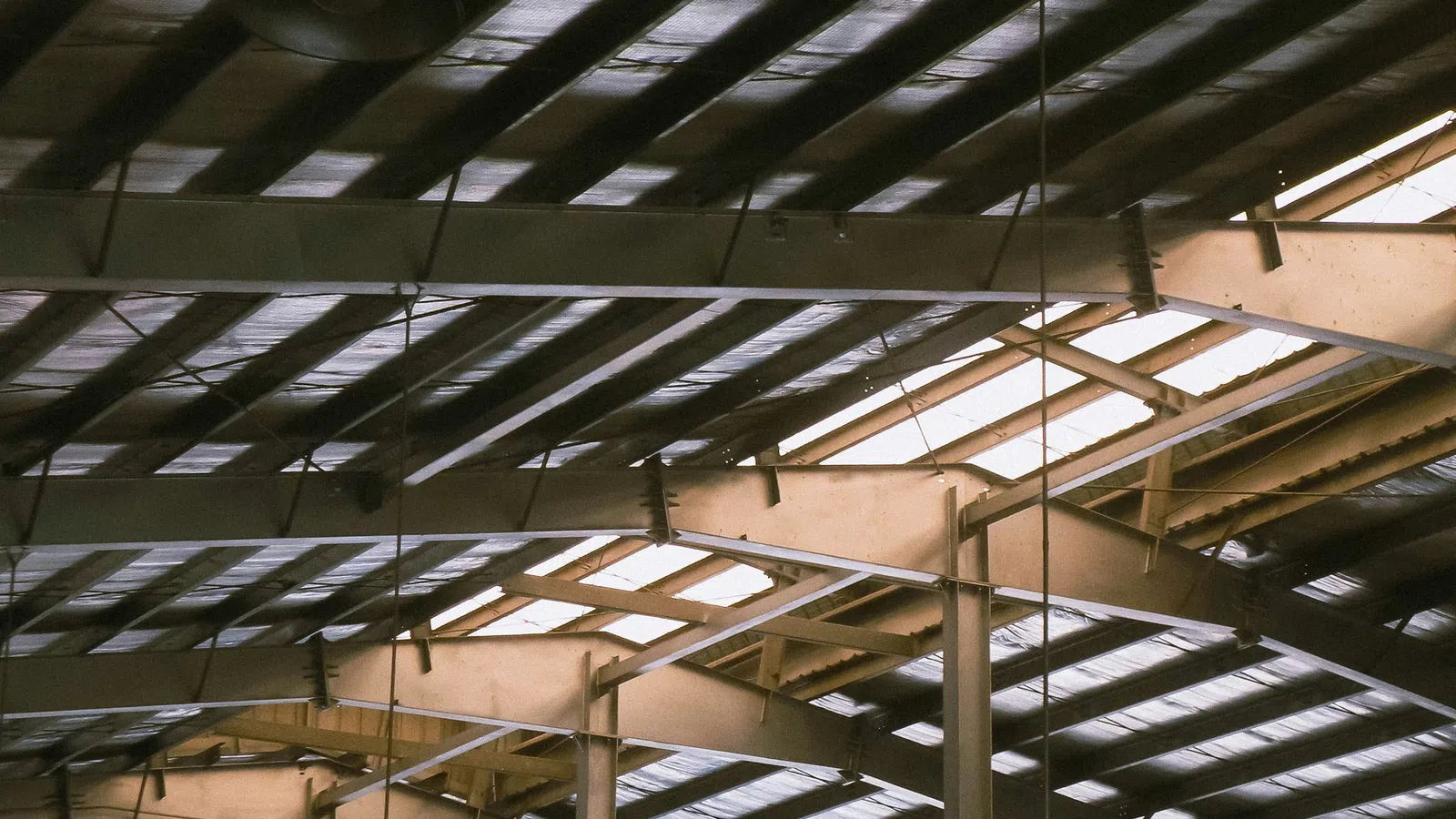 Interior view of a warehouse ceiling with metal beams and hanging industrial lights during daytime.