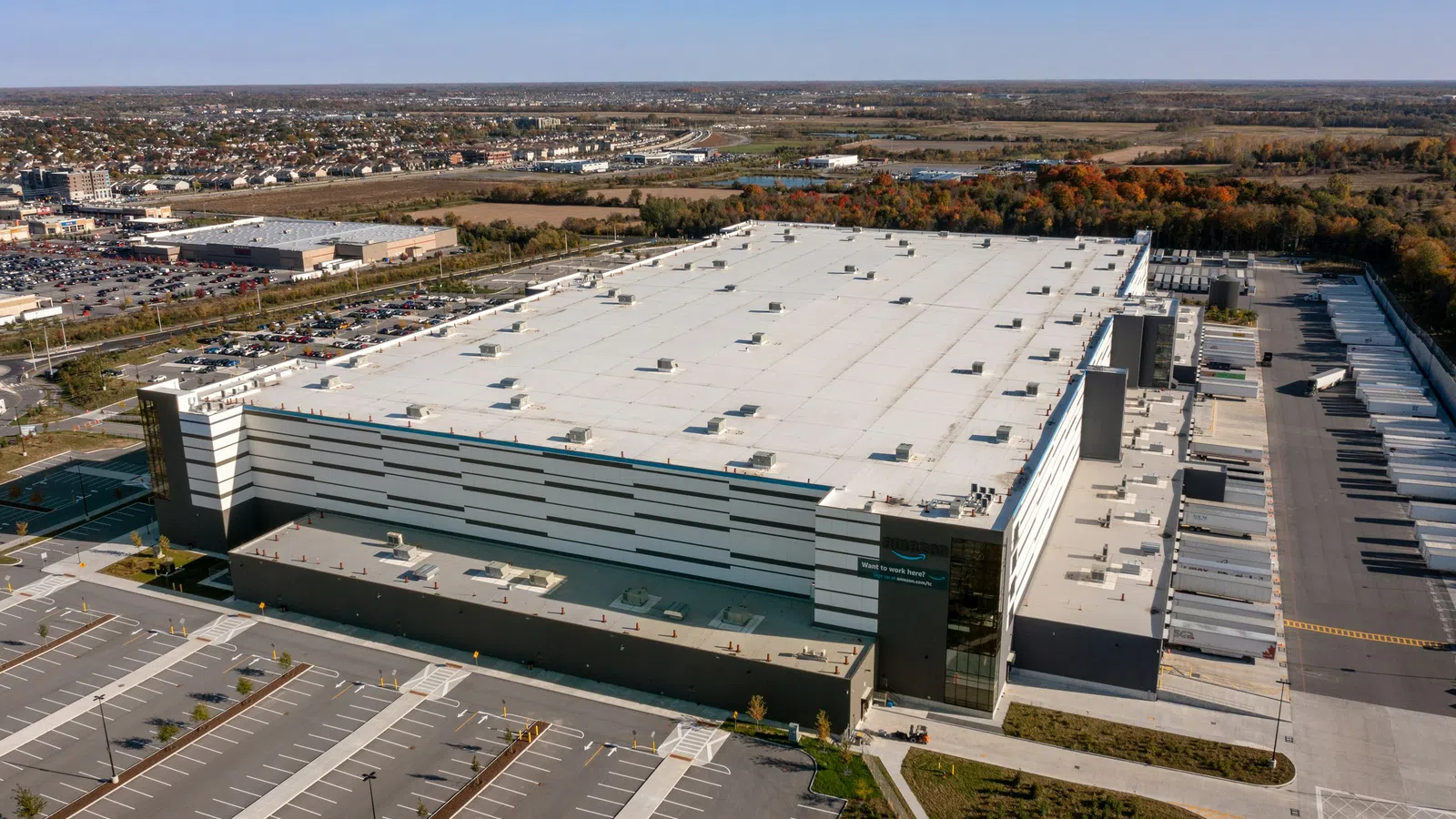 Aerial view of a large industrial warehouse with multiple loading docks and an adjacent empty parking lot under clear blue sky.
