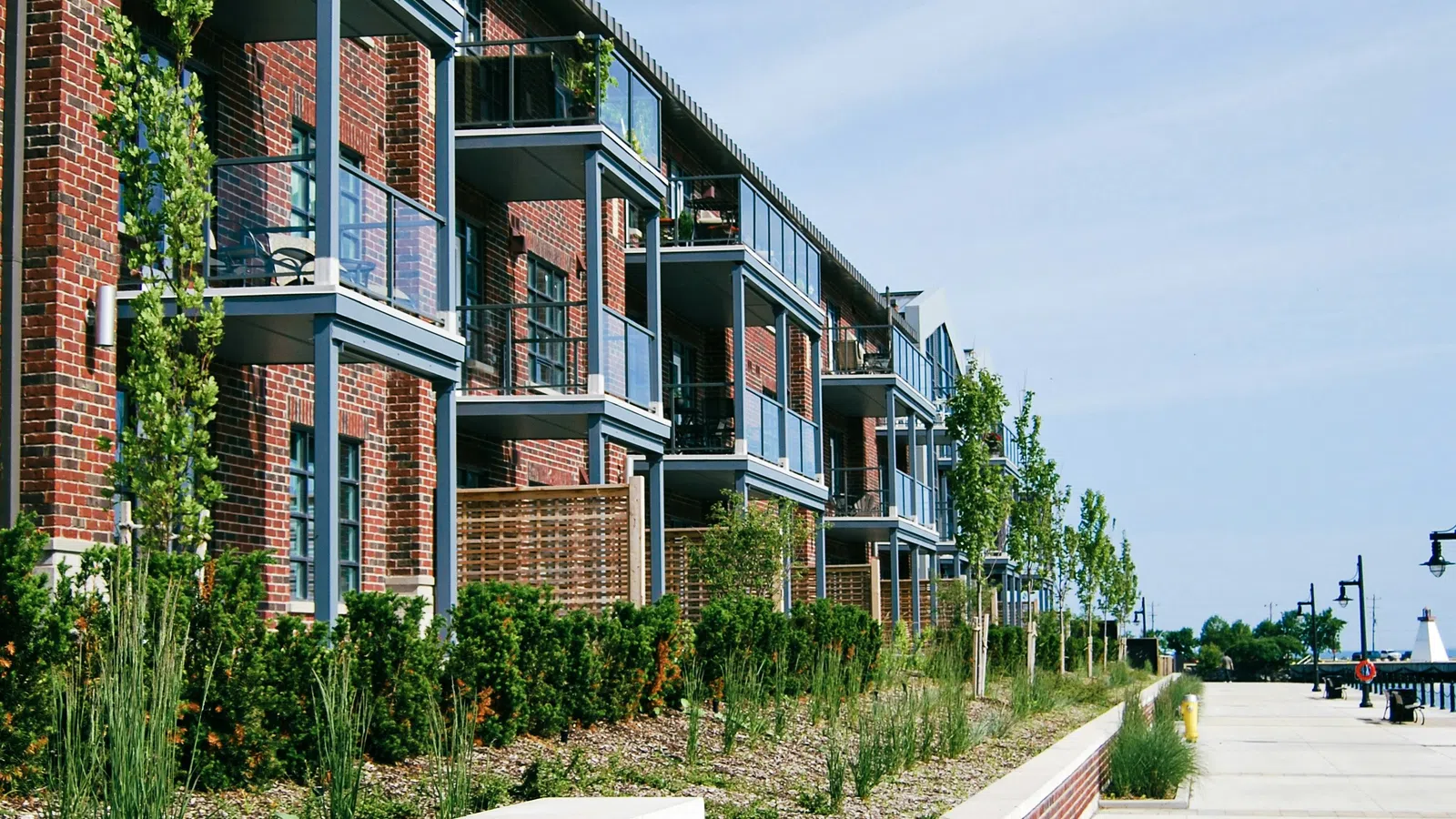 Modern brick apartment building with glass balconies and landscaped sidewalk on a sunny day.