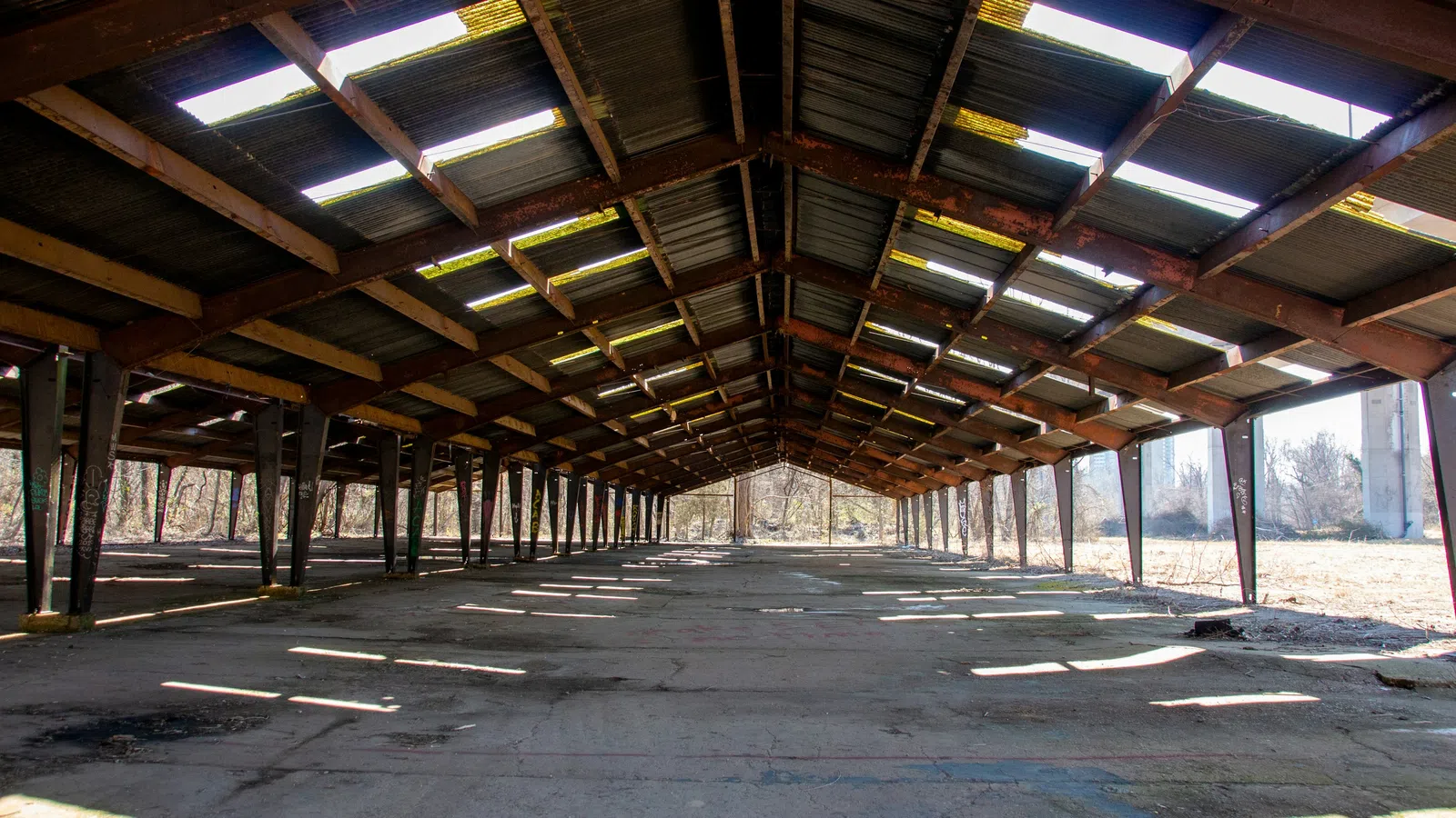 Empty abandoned warehouse with rusted metal beams and concrete floor under a partially covered roof.