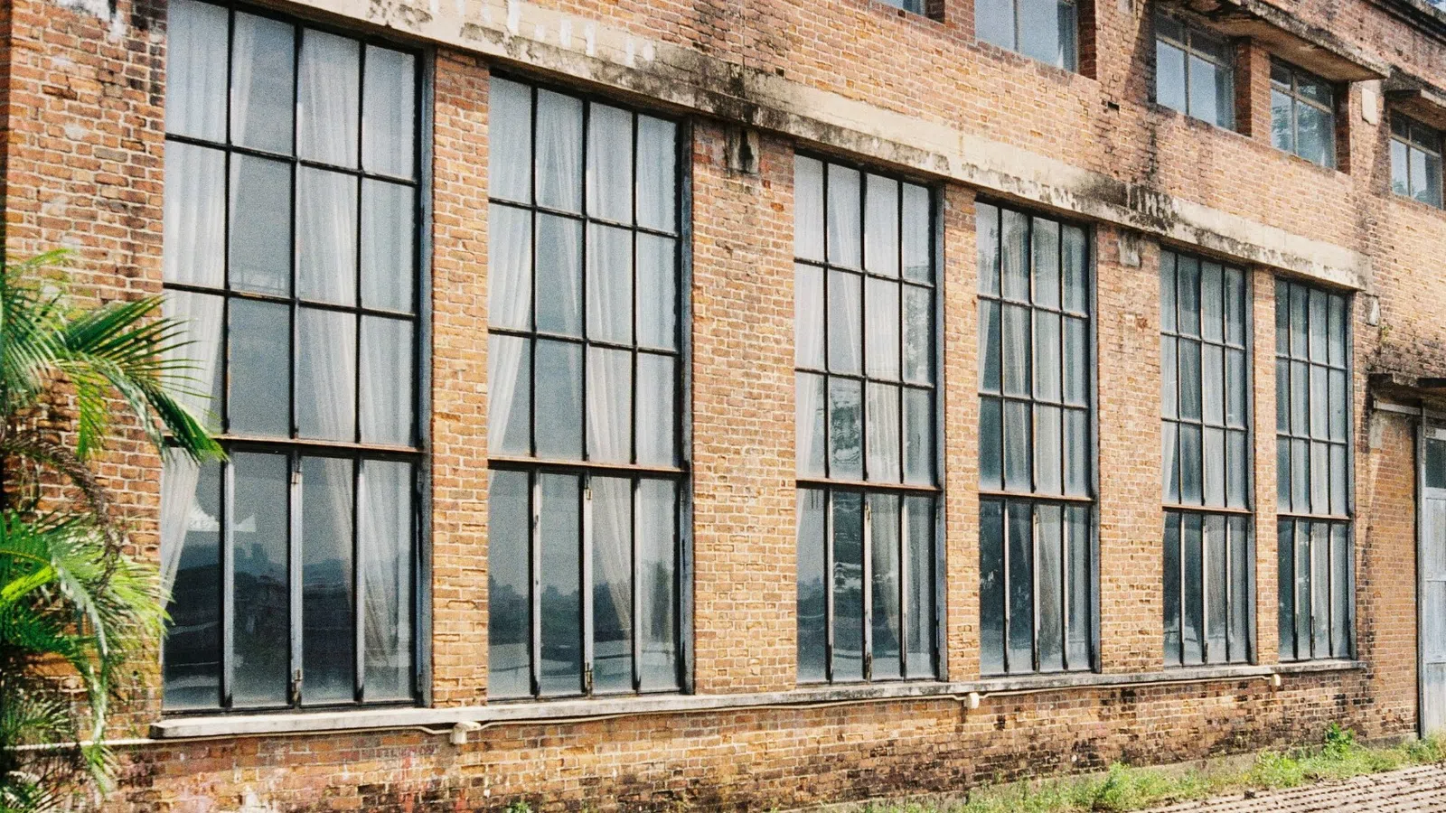 Construction worker in safety vest and helmet inspecting industrial building with large windows and brick walls.