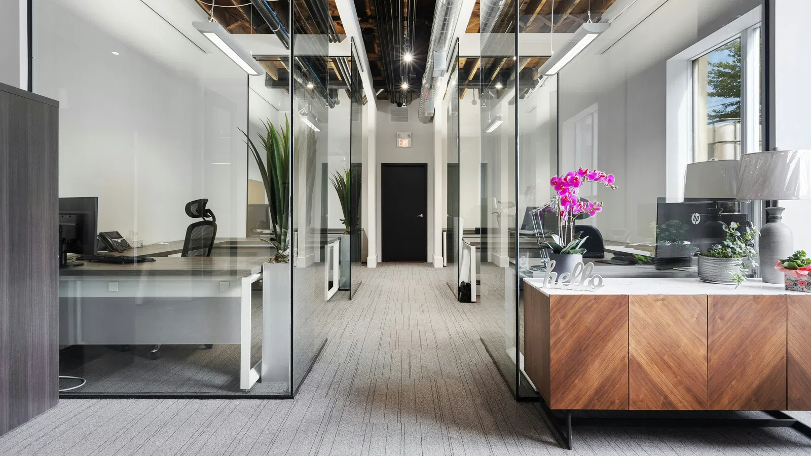 Modern office hallway with glass-walled workspaces, wood cabinets, plants, and exposed ceiling ducts under bright lights