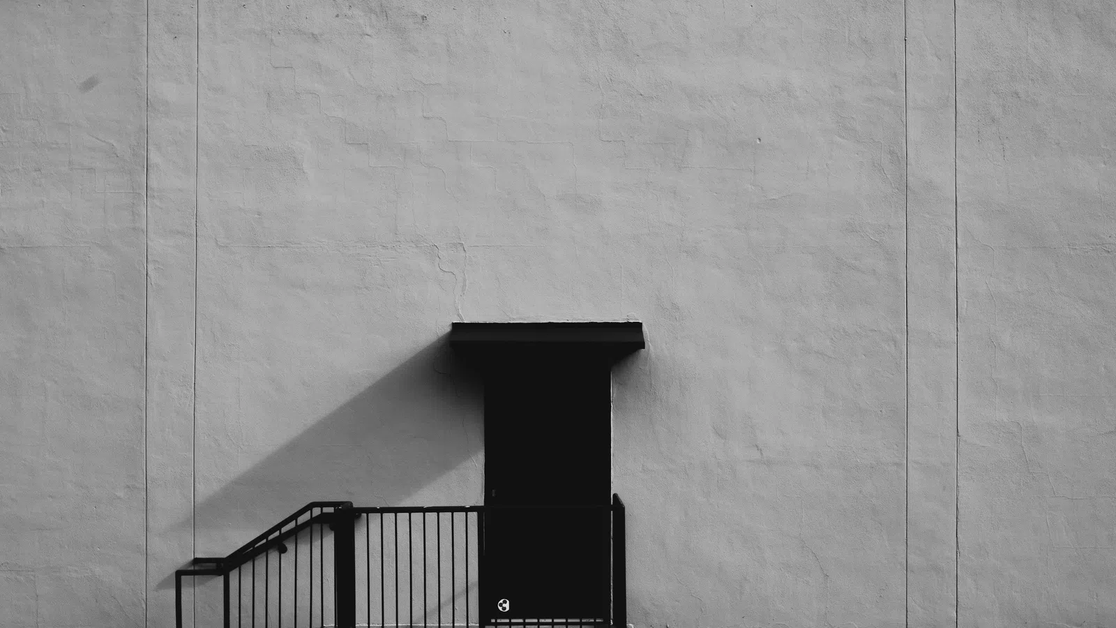 Black door with shadow and metal railing on plain light gray textured wall in minimal urban setting