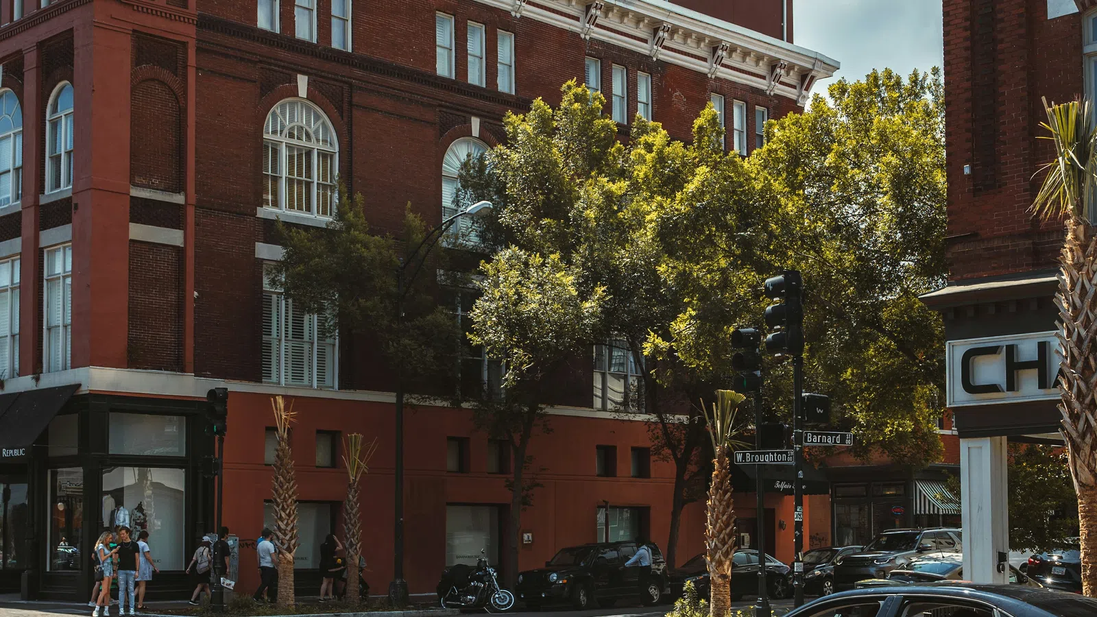 Sunny urban street scene with red brick buildings, cars parked, and pedestrians crossing at the intersection.