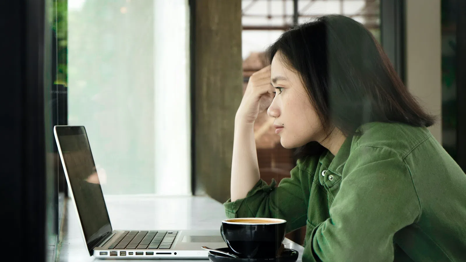 Young woman in green jacket intently focused on laptop screen with coffee cup in front indoors