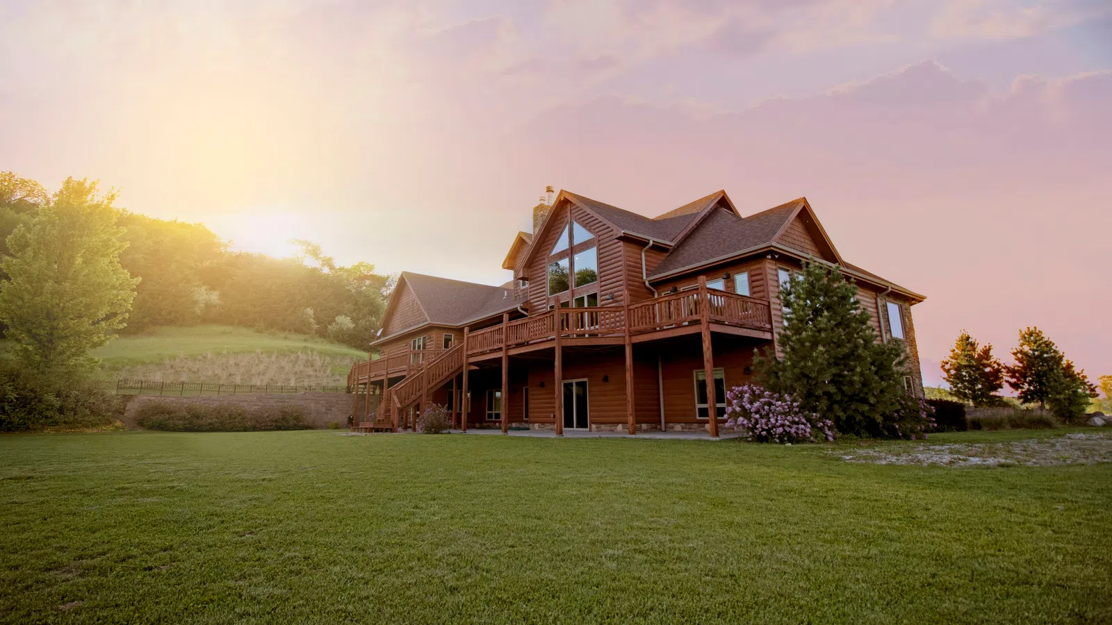 Spacious wooden cabin with large windows and wraparound deck surrounded by greenery at sunset.