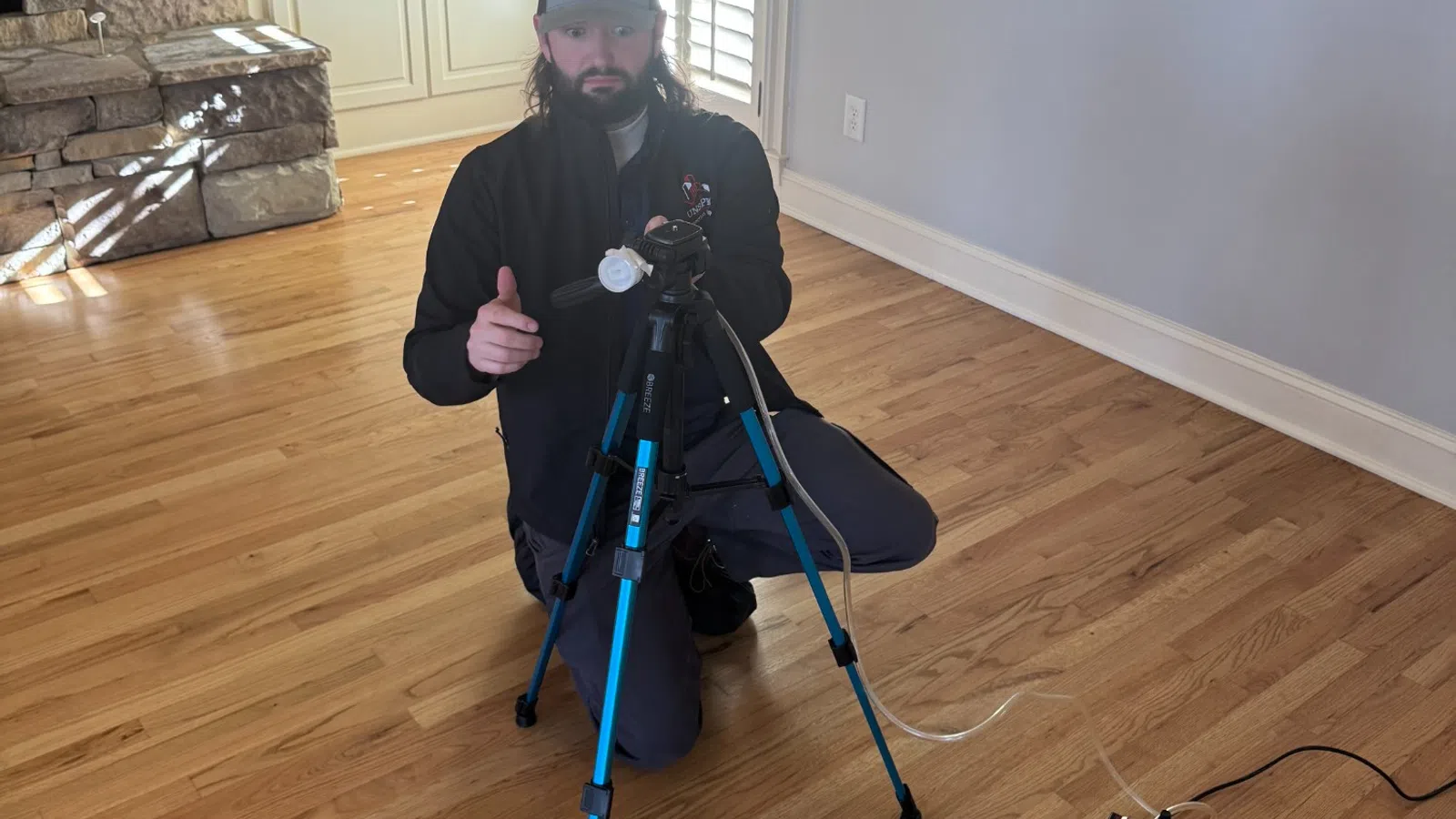 Man kneeling on hardwood floor with tripod and surveying equipment in a bright room with stone fireplace.