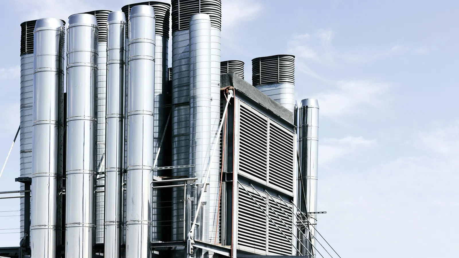 Shiny silver industrial ventilation pipes and chimneys against a clear blue sky in a factory setting.