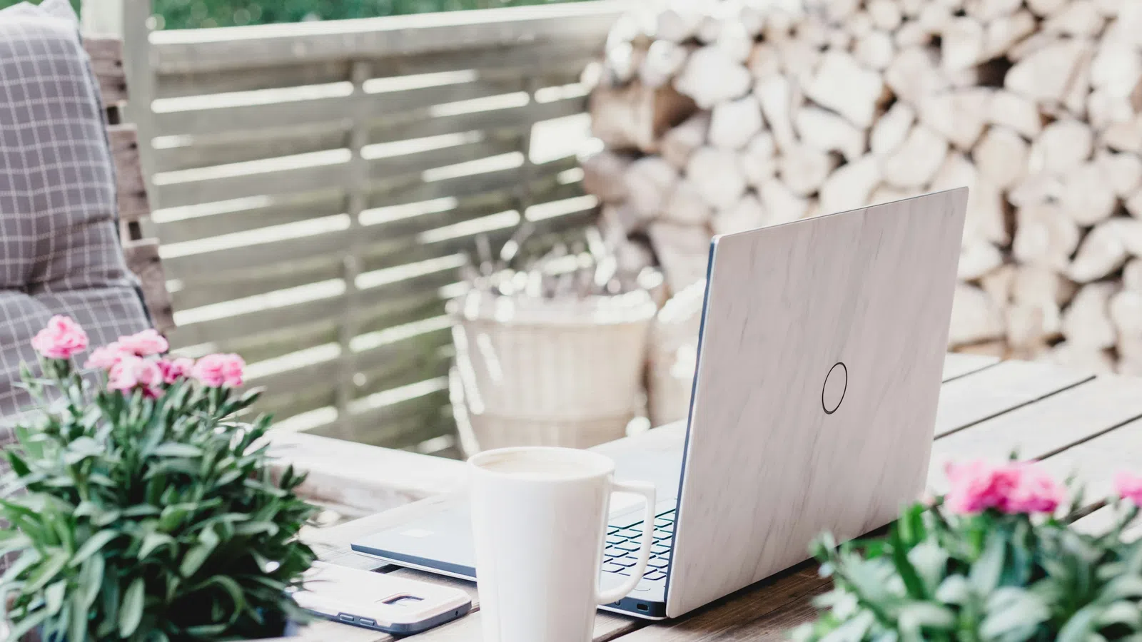 Cozy outdoor workspace with laptop, coffee mug, smartphone, and potted flowers on a rustic wooden table.