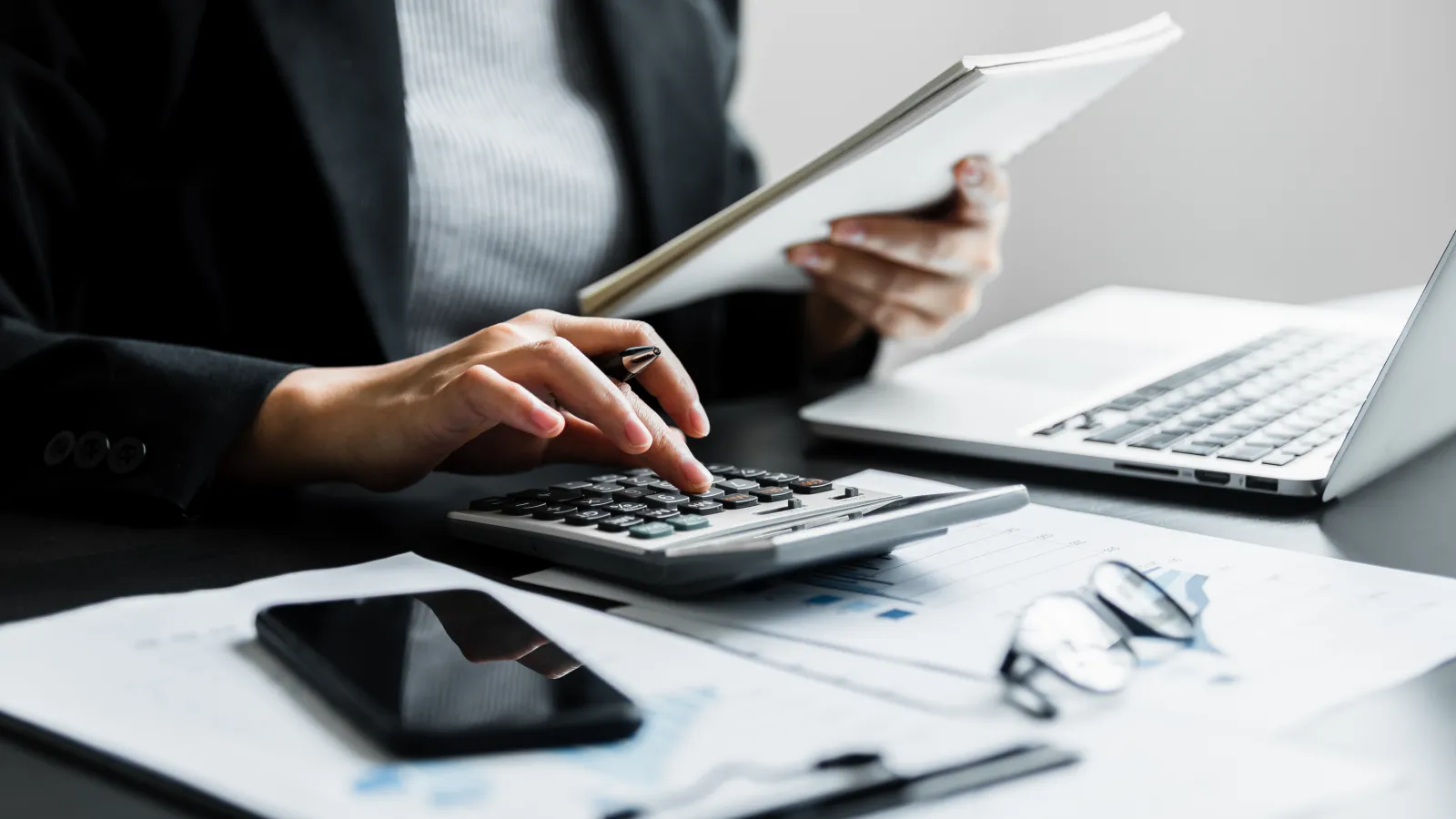 Person using calculator and holding documents at desk with laptop, phone, and glasses on papers.