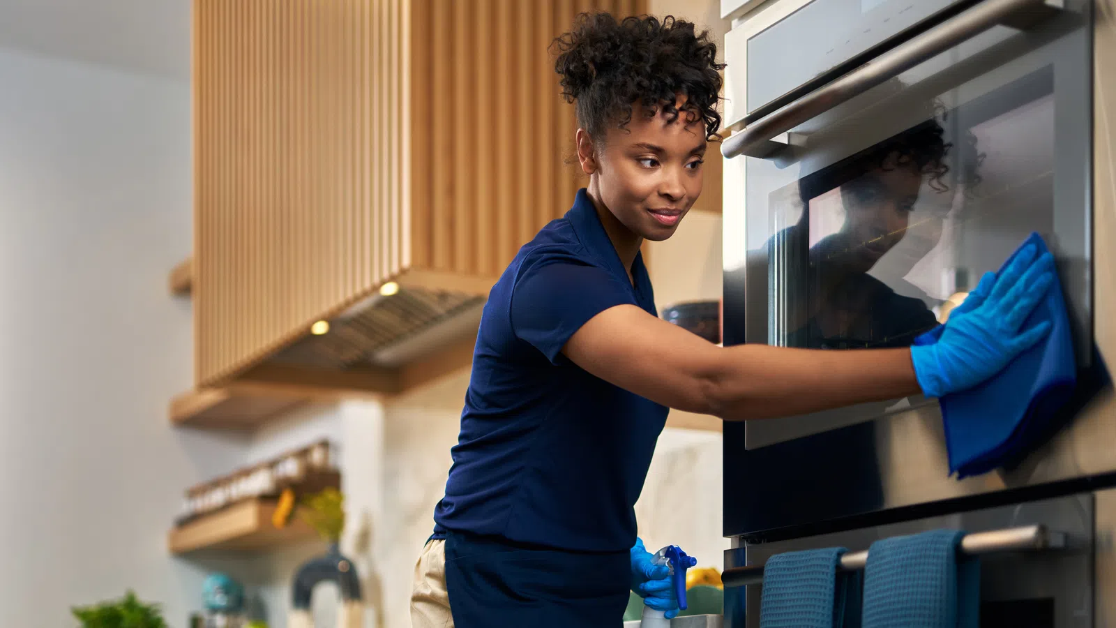 Woman in blue gloves cleaning the stainless steel oven door in a modern kitchen with a spray bottle and cloth.