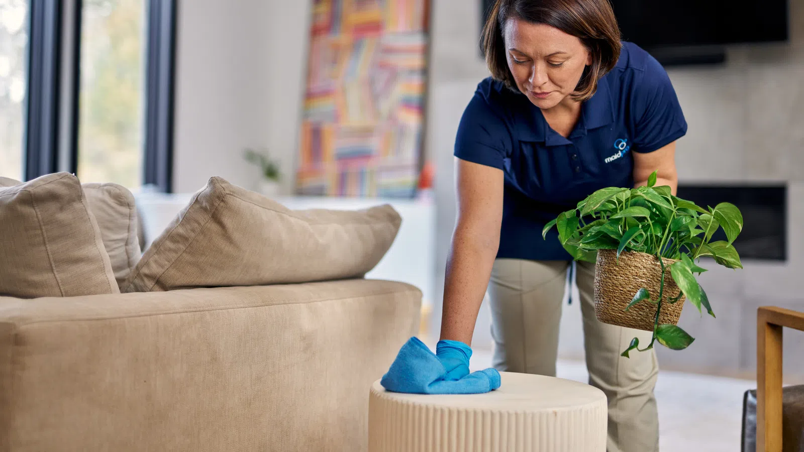 Woman cleaning a beige ottoman with a blue cloth while holding a green potted plant in a modern living room